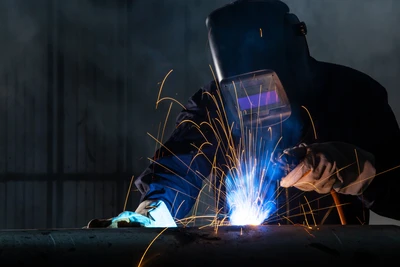 Soudeur professionnel en action dans un atelier industriel, portant masque de protection et combinaison bleue, effectuant une soudure à l'arc avec projections d'étincelles dorées et flamme bleue intense